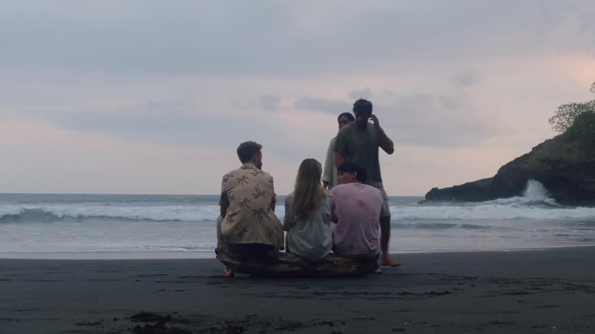 A group of diverse friends gathers on a dark sand beach at dusk, sitting on driftwood and watching the ocean waves roll in