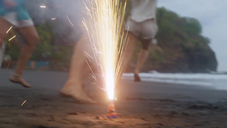 Friends light a fountain firework on a black sand beach and run away in slow motion as one of them does a backflip