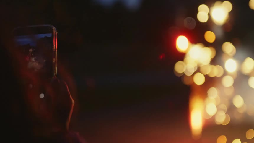 A person films a sparkling firework fountain with their mobile phone during a nighttime celebration