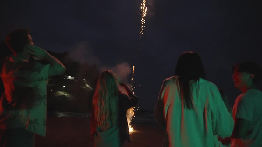 A diverse group of young friends celebrate on the beach at night, watching fireworks explode in the dark sky above