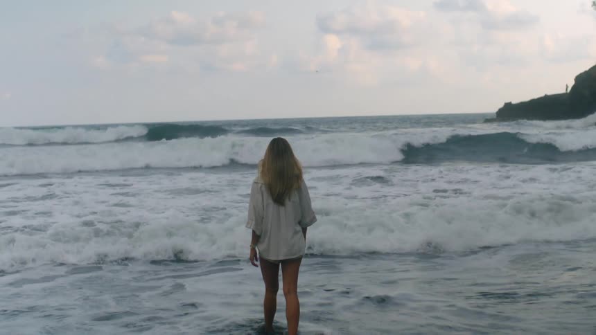 A woman with long blonde hair walks into the powerful, foamy ocean waves in slow motion