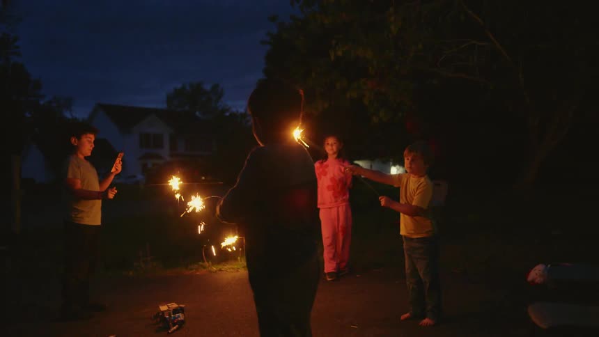 A group of young children celebrate in slow motion by waving bright sparklers in their neighborhood at dusk