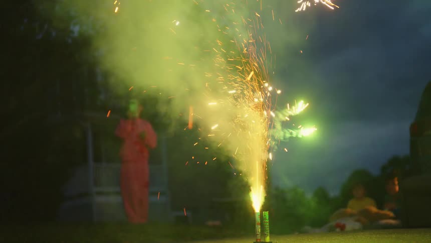 A ground fountain firework erupts in a shower of colorful sparks and smoke on a suburban driveway at dusk while a family watches