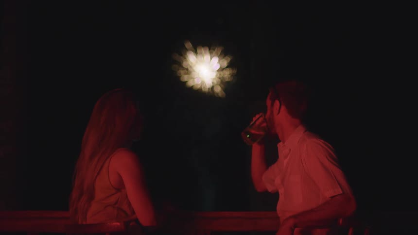 A couple enjoys a romantic evening with drinks, talking and watching a stunning fireworks display from a balcony