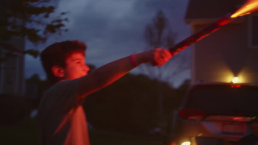 Slow motion close-up of a young boy holding a firework at dusk as colorful sparks illuminate his face with wonder