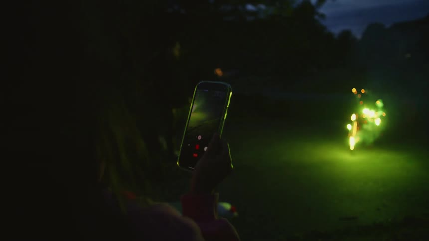 Over the shoulder shot of a person recording a vibrant green firework exploding on the ground at night with their smartphone