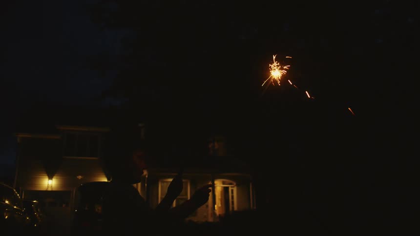 A person's silhouette is visible as they wave a bright sparkler in slow motion during a nighttime celebration