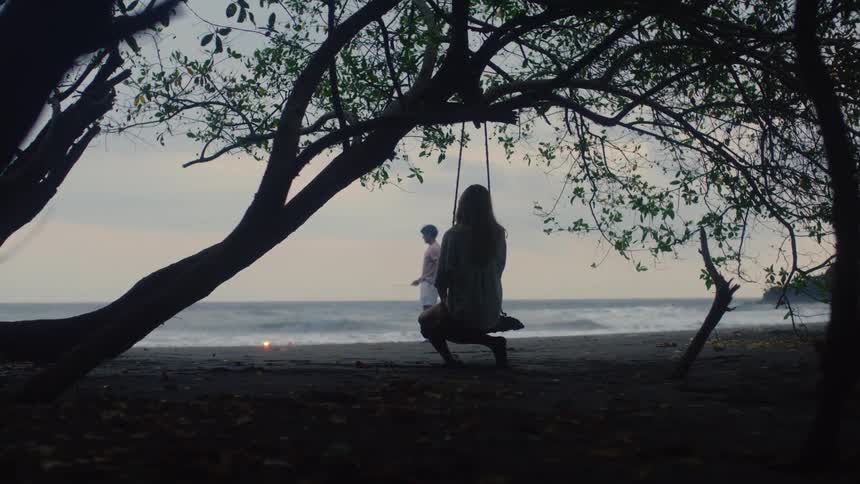 A woman on a tree swing watches a man light a sparkler on a dark sand beach at dusk