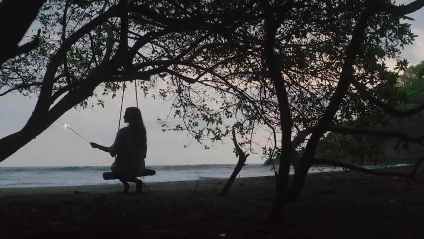 Silhouetted woman on a tree swing holds a sparkler and looks out at the ocean on a dark sand beach
