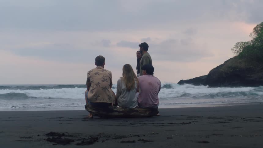 Friends sit on a log on a dark sand beach watching the waves while another couple walks past