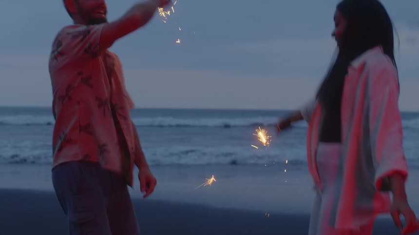 A joyful couple celebrates on a dark beach at dusk, dancing and waving lit sparklers with the ocean waves behind them