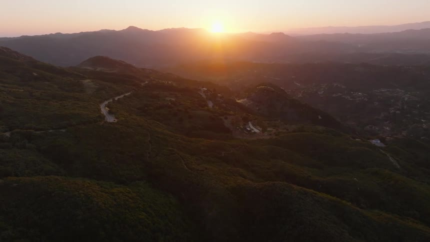 A serene aerial view shows the sun rising over a lush, green mountain range with a winding road