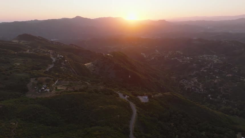 A serene aerial view captures the sun setting over a vast, green mountain range with a winding road cutting through the hills