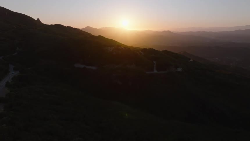 Slowly flying over a scenic mountain pass with a winding road as the golden sun sets in the distance