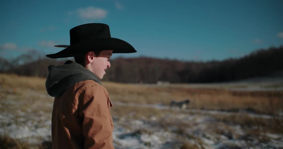 A young boy in a cowboy hat watches his dog walk across a snowy, sunlit pasture on a crisp winter day