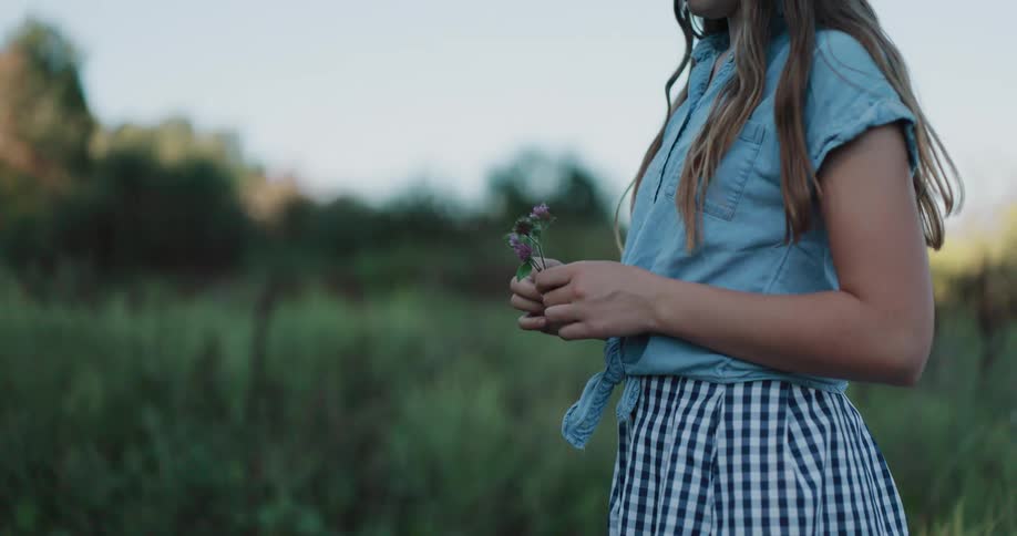A young woman wearing a denim shirt and gingham skirt stands in a grassy field holding a small purple wildflower