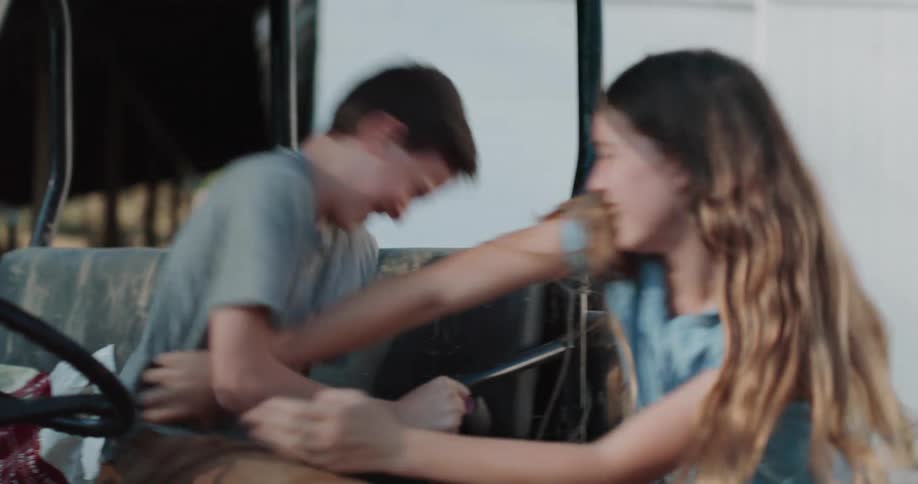 A sister playfully tickles her laughing brother as he sits in the driver's seat of a utility vehicle on a farm