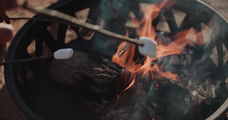 Slow motion close-up of two marshmallows on sticks being toasted over the flames of a campfire in a fire pit