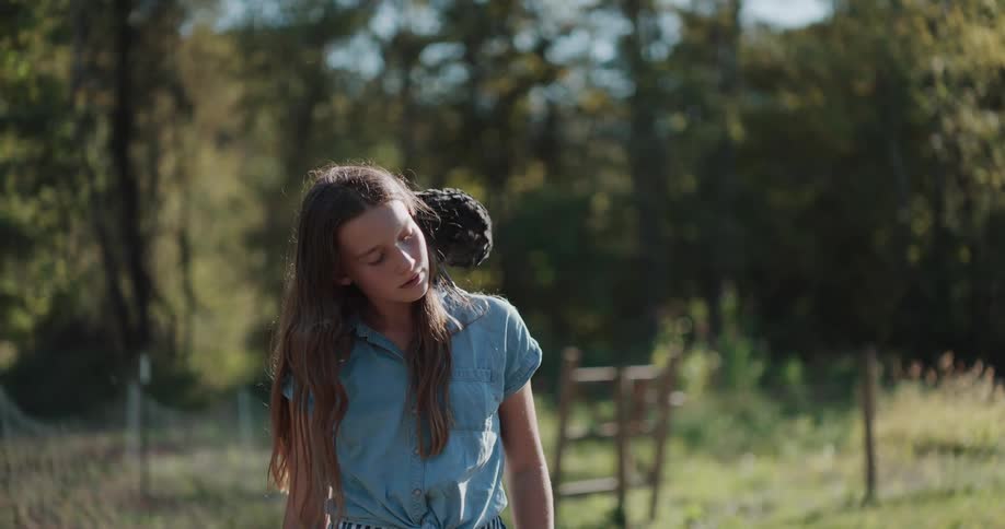 A young girl gently holds and pets a unique Polish crested chicken perched on her shoulder in a sunlit country field
