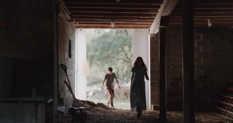 Large white barn doors slowly open to reveal a girl and boy who walk from the sunlit outdoors into the dark barn