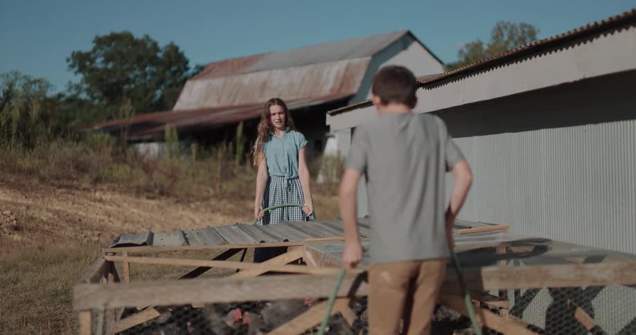 A teenage girl and boy work together in slow motion to move a mobile chicken coop across the pasture on their farm