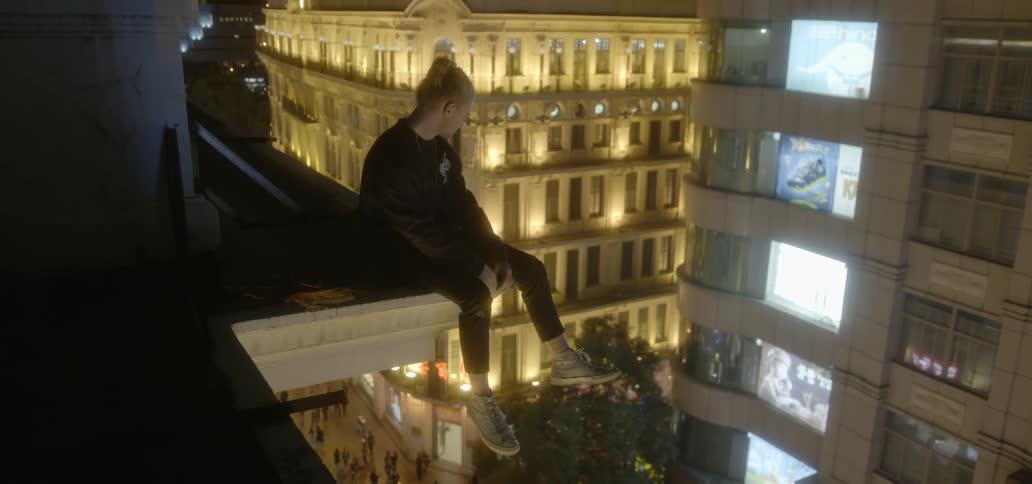 A young man sits on a rooftop ledge overlooking a city at night