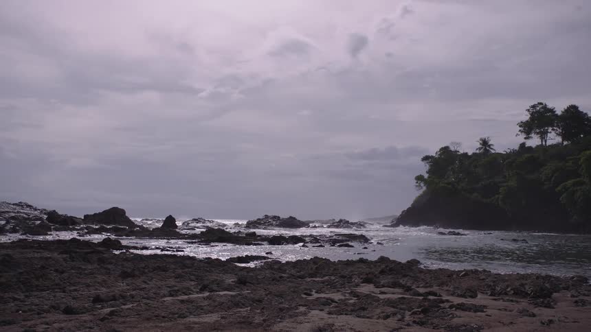 A moody seascape shows gentle waves washing over a rocky shoreline under a cloudy sky