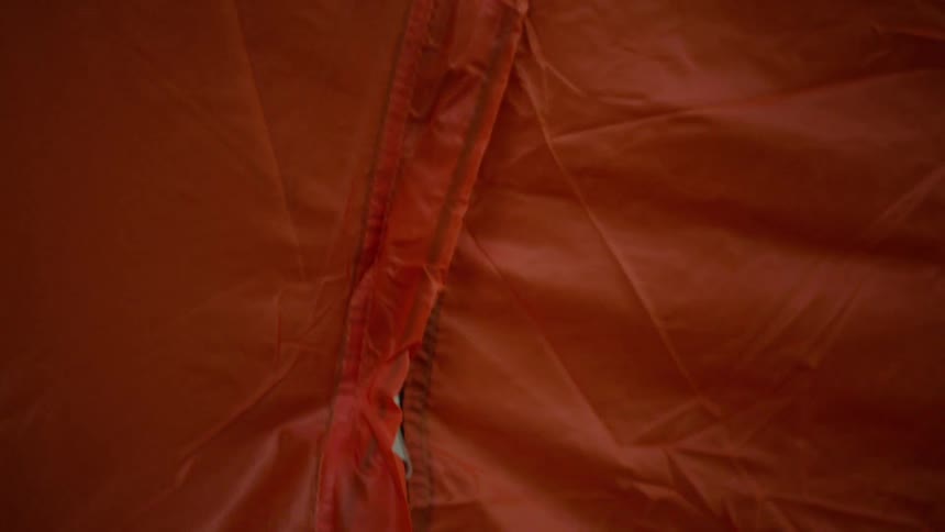 A close-up shot from inside a tent shows a hand zipping the entrance shut for the night