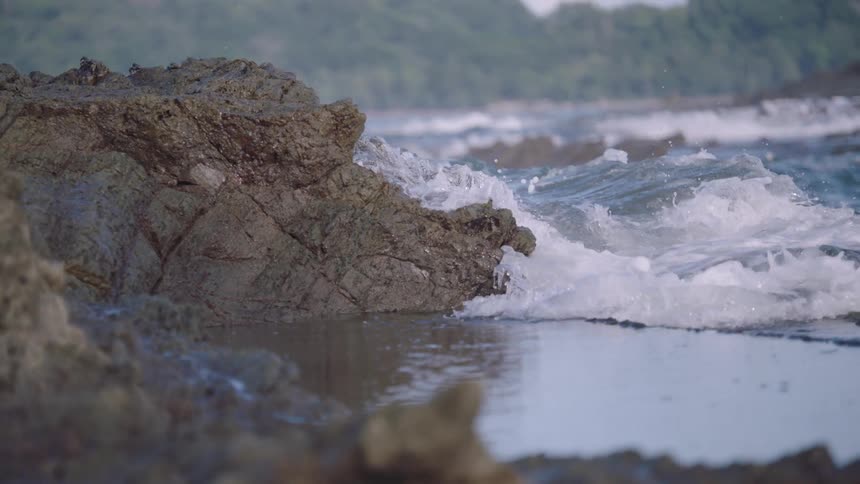 A close-up slow-motion shot of ocean waves crashing against rugged coastal rocks, with water splashing and receding into a tide pool