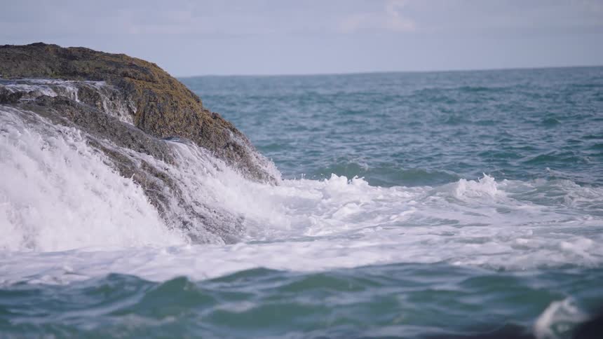 Slow-motion close-up of powerful ocean waves crashing over a rocky shoreline under a cloudy sky