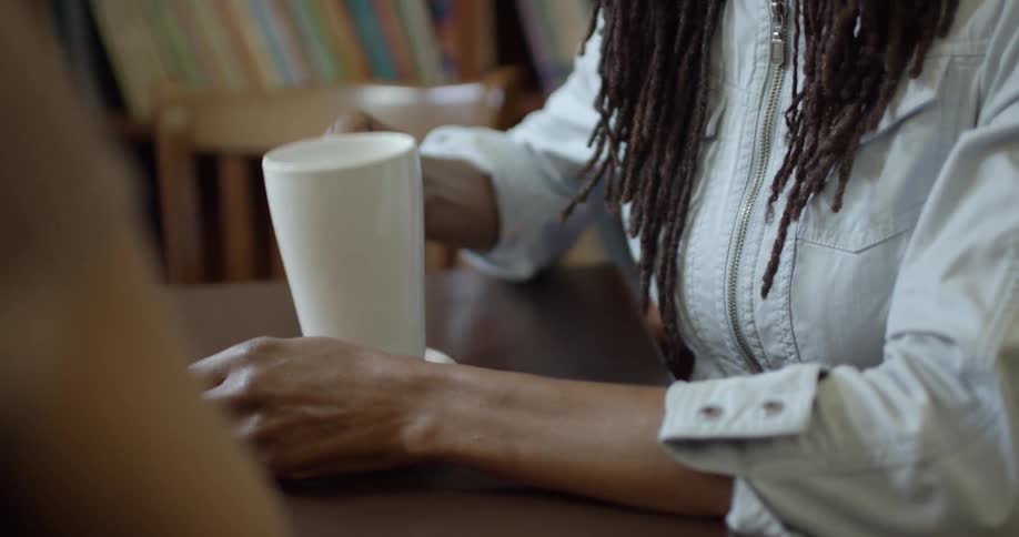 Woman sitting next to another person at table in library takes drink from mug