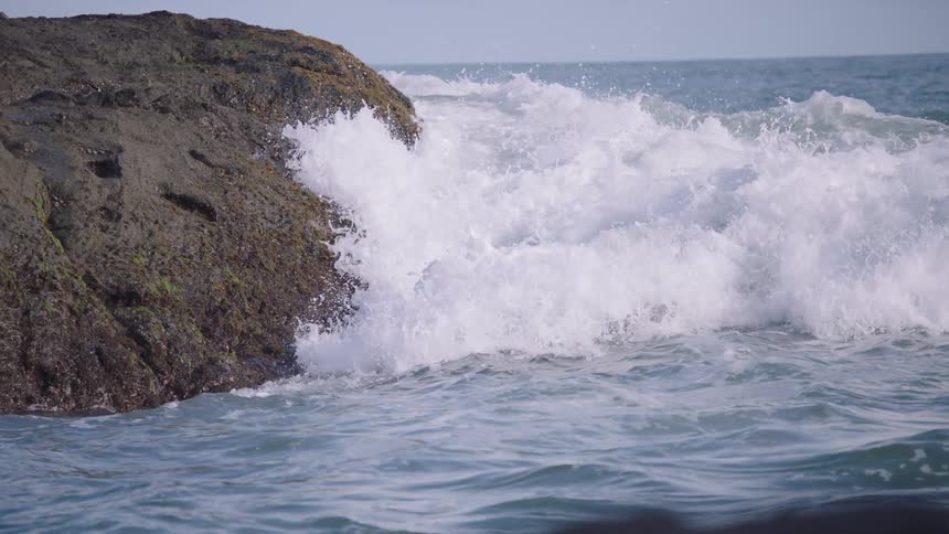 Powerful ocean waves crash against a moss-covered rock in dramatic slow motion