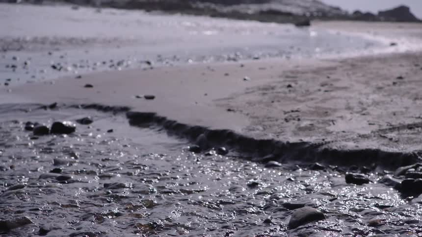 A gentle stream flows over pebbles and sand, carving a path towards the calm ocean in the background