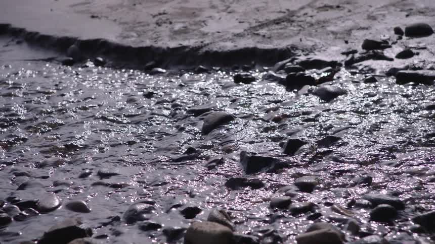 A peaceful slow-motion close-up of a shallow stream shimmering as it flows over smooth rocks on a sandy bank