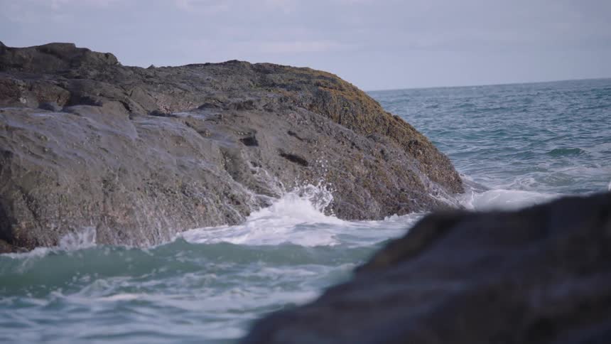 A slow-motion close-up of ocean waves crashing against a large rock on a rugged coastline