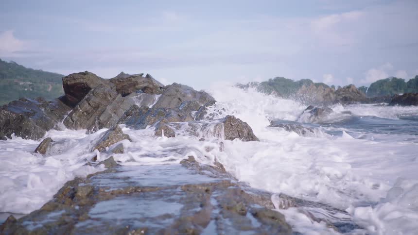 Slow-motion footage of powerful ocean waves crashing over a rocky shoreline, creating white foam
