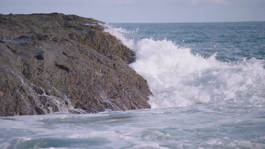 Powerful ocean waves crash against a large coastal rock in dramatic slow motion