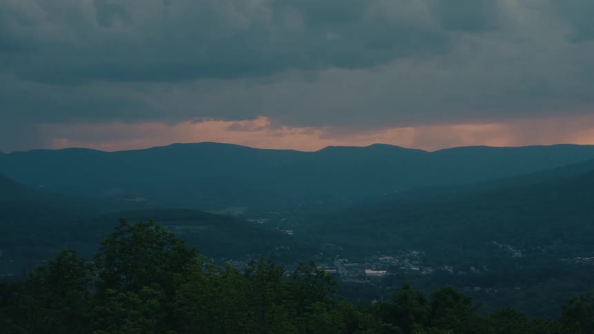 A wide, dramatic view of a small town nestled in a mountain valley under a stormy sky at sunset