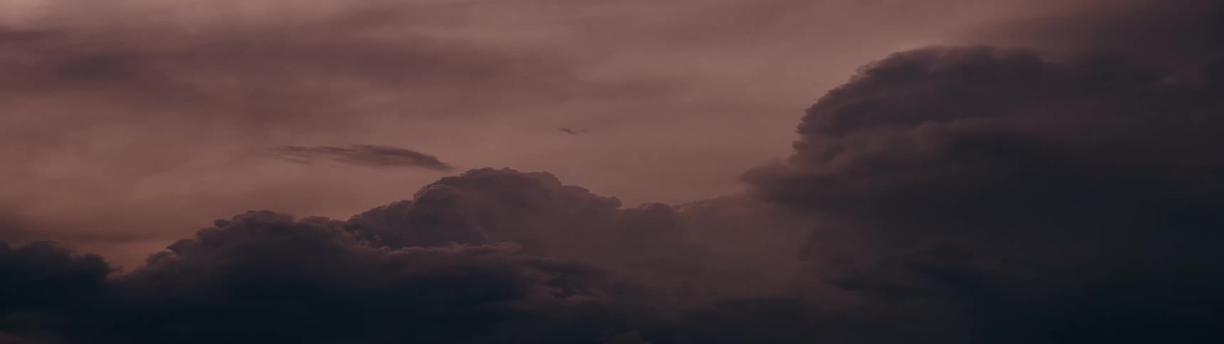 A lone airplane flies across a vast sky filled with dark, moody clouds at dusk