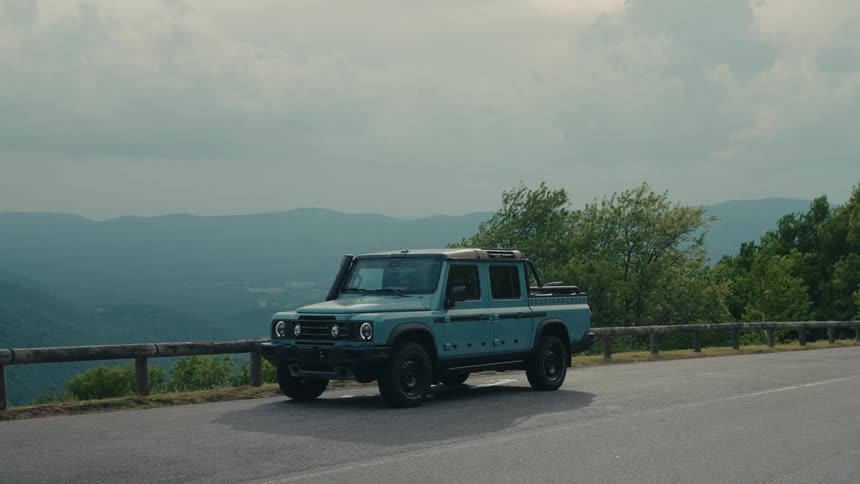 A light blue off-road truck is parked on a scenic mountain road overlooking a vast, hilly landscape