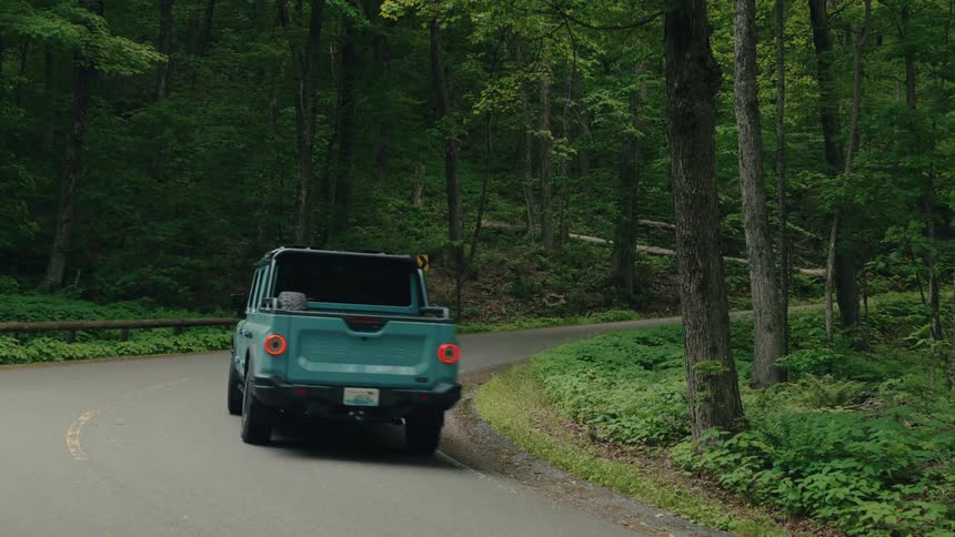 A teal pickup truck drives away down a scenic winding road through a lush green forest