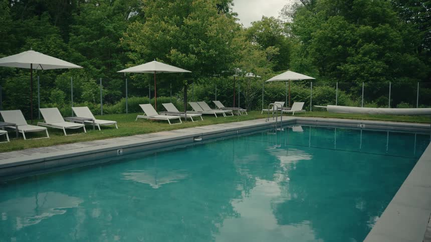 A quiet, still shot of an empty outdoor swimming pool with lounge chairs and umbrellas set up on the grass under an overcast sky