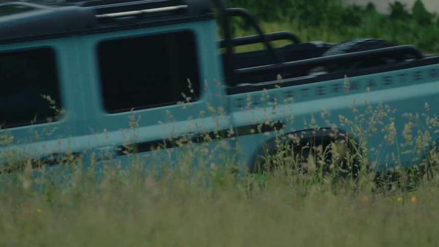 A teal off-road pickup truck with its headlights on drives through a field of tall green grass from right to left