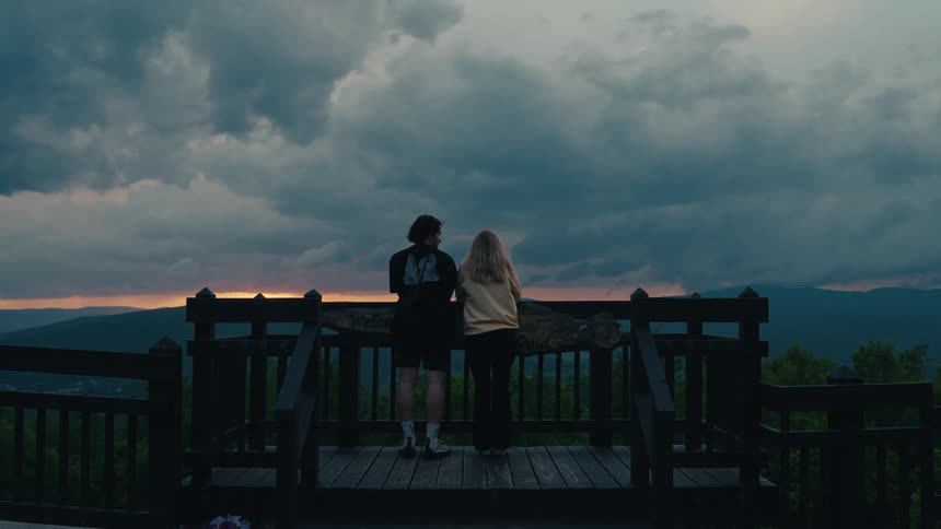 A couple stands together on a wooden viewing platform, watching a dramatic sunset over rolling mountain ranges