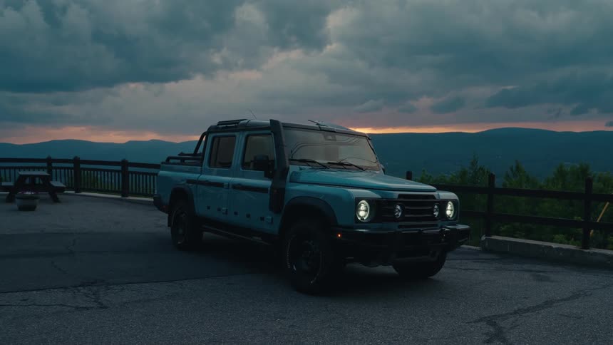 An off-road pickup truck is parked at a scenic mountain overlook with its headlights on under a dramatic twilight sky