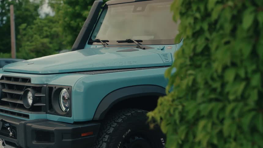 A static close-up shot of the front of a modern teal Ineos Grenadier SUV parked outdoors with green foliage in the background