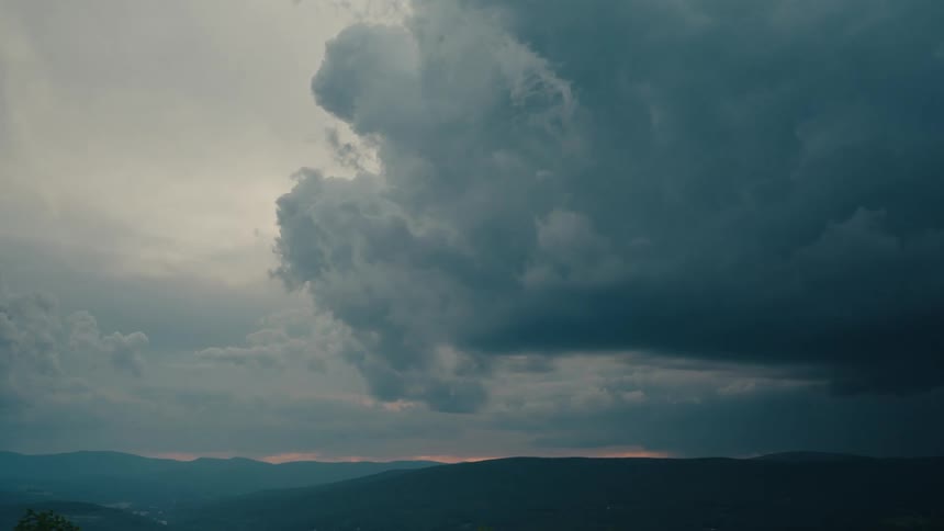 A dramatic, dark storm cloud looms over a mountain range as the sun sets on the horizon
