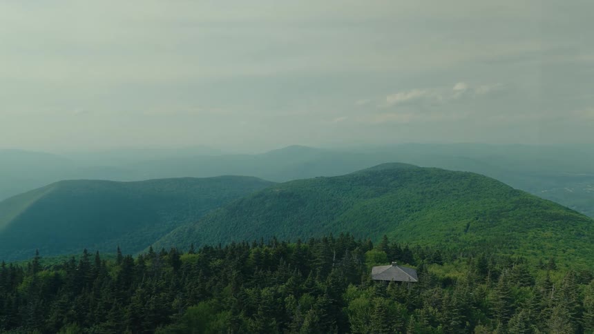 A serene, hazy view from a mountaintop reveals a small shelter nestled in the lush green forest below