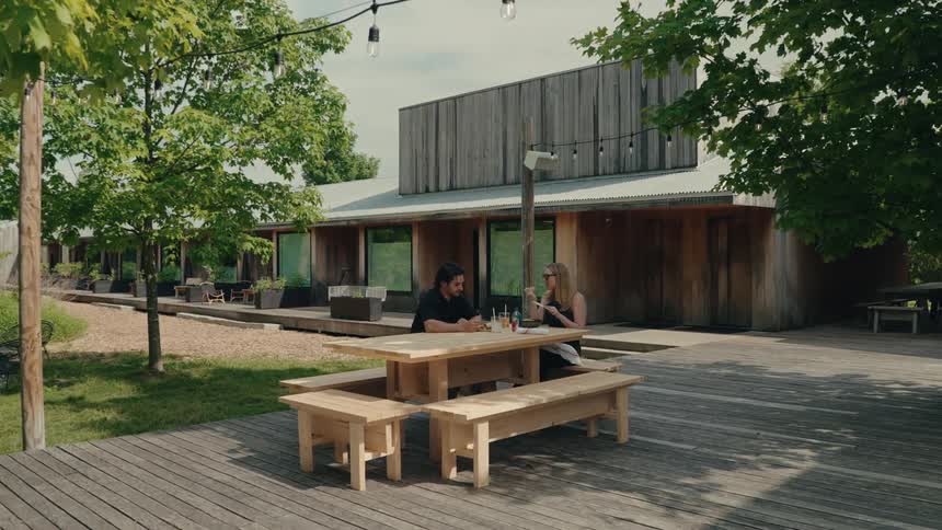 A couple sits and talks at a wooden picnic table on the deck of a modern rustic home on a sunny day