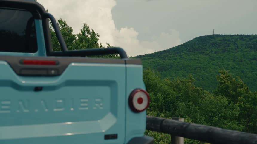 Rear close-up of a light blue off-road truck parked at a scenic viewpoint overlooking a windy, forested mountain